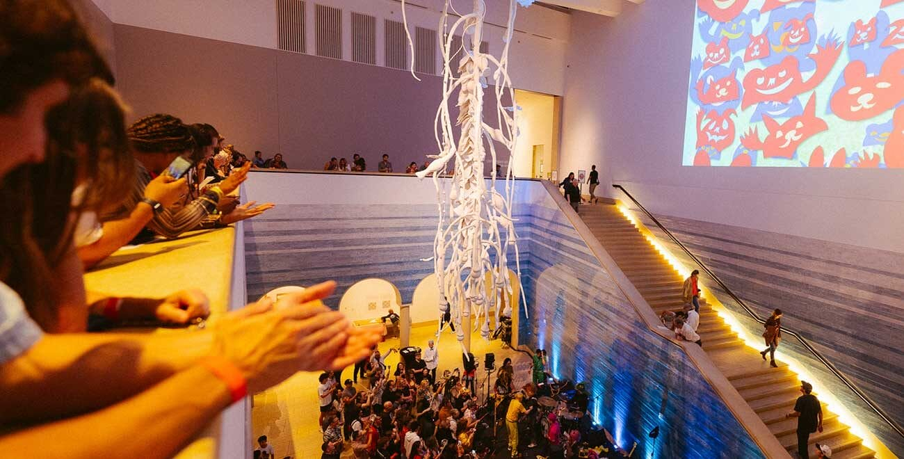 An indoor event space with a large staircase leading down to a lower level. People are on both levels, with some on the upper balcony, clapping and using their phones. A large white hanging sculpture is in the middle of the space. The wall to the right has a projection of colorful, cartoon-like animal faces. The lower level has a crowd of people and some musical instruments, suggesting a gathering or performance. The space is well-lit with warm lighting along the staircase.