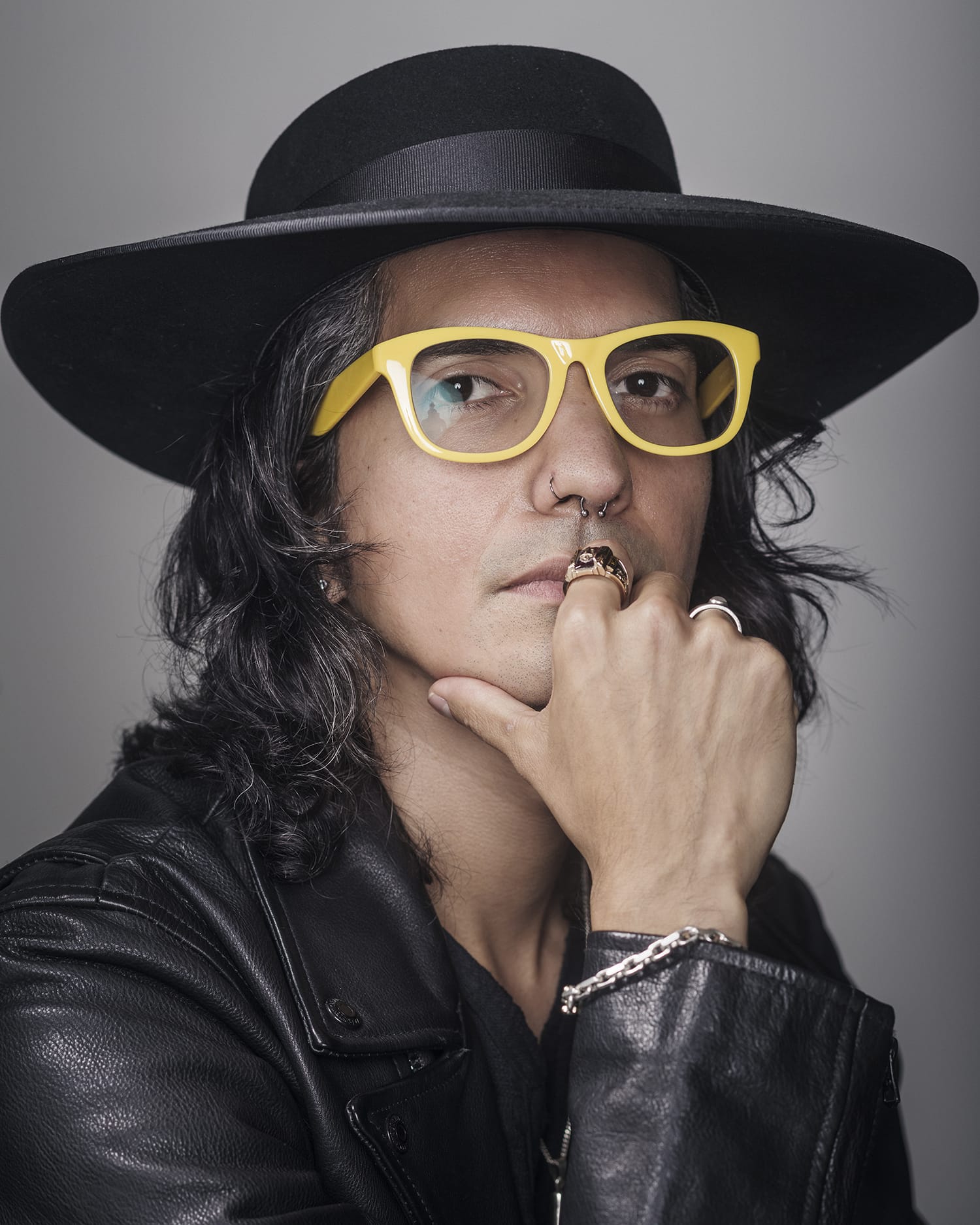 A headshot of a person wearing a black wide-brimmed hat, yellow rimmed glasses and a black jacket, with his hand on his chin